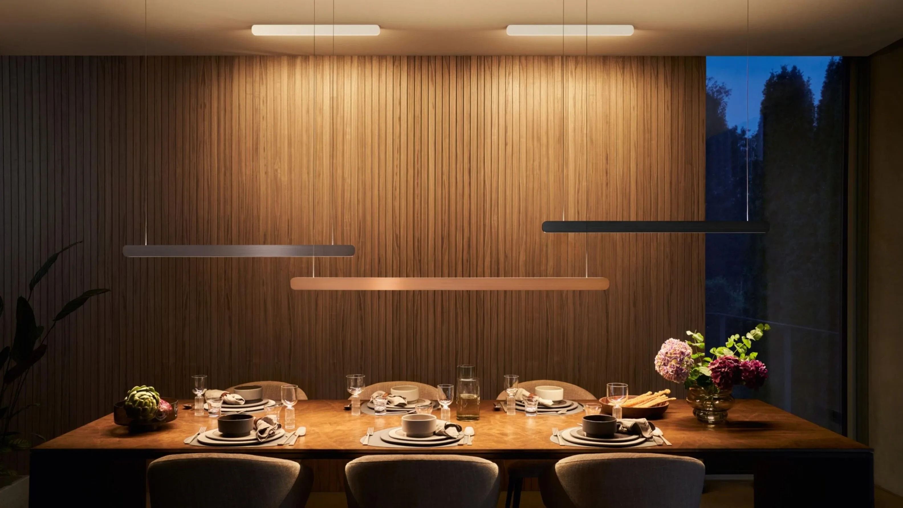 Three slim linear pendants staggered over a dining table against timber battens